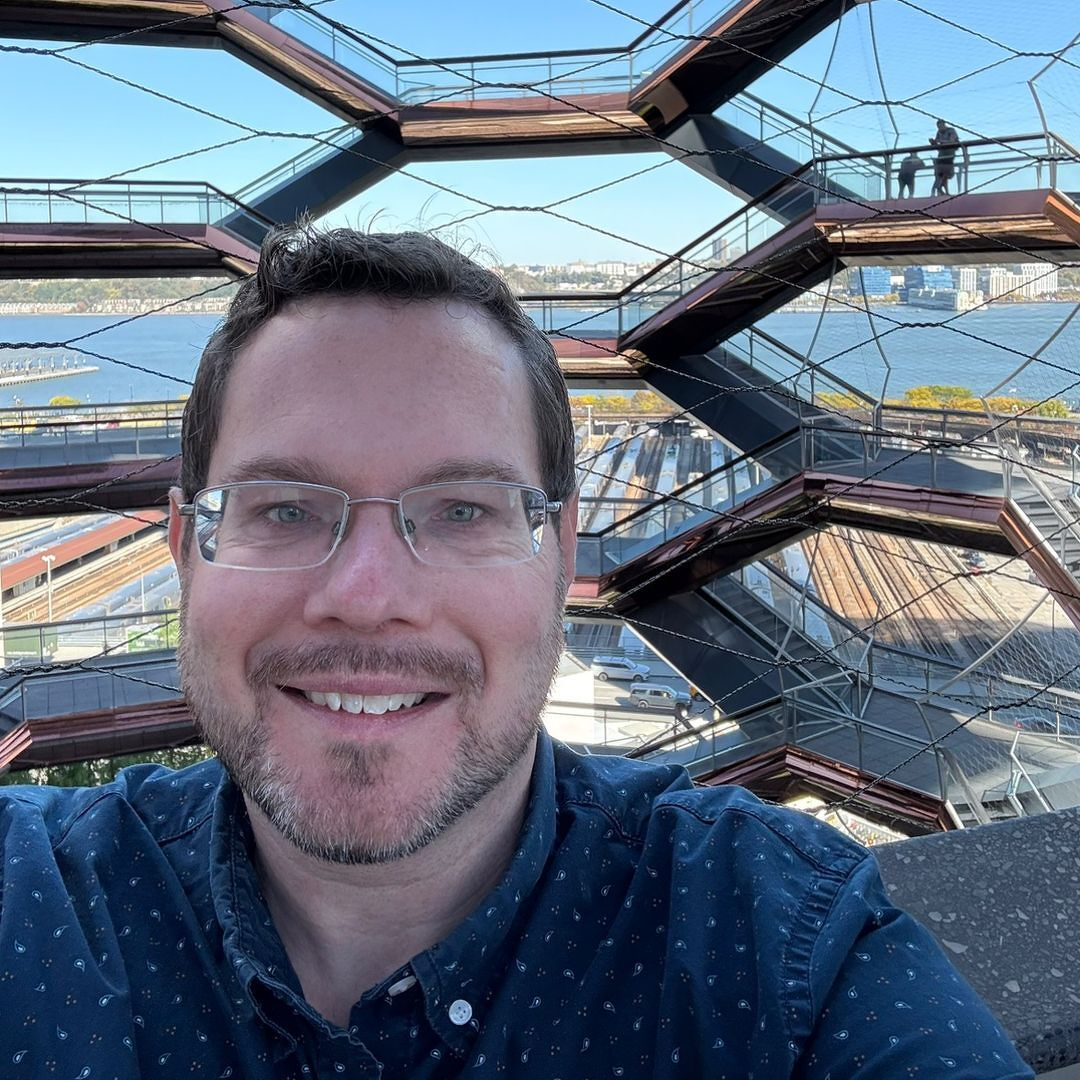 A smiling man wearing glasses takes a selfie with the intricate stair structure of the Vessel and waterfront in the background.